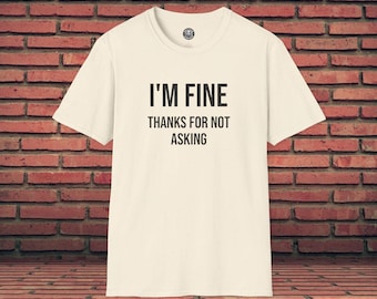 May include: Cream-colored t-shirt with the text "I'M FINE THANKS FOR NOT ASKING" in black. The shirt has a crew neck and short sleeves. The background is a brick wall.