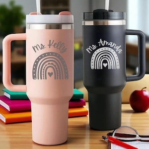 May include: Two insulated tumblers, one blush pink and one black, with handles and lids. Each tumbler features a rainbow design and the name "Ms. Kelly" or "Ms. Amanda". The tumblers have a straw and are designed for cold beverages.