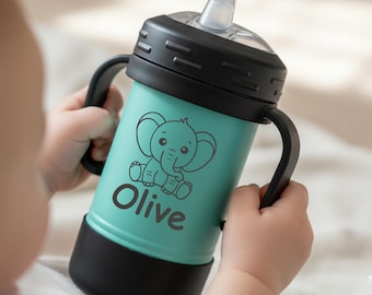 May include: A teal baby sippy cup with black accents and handles. The cup features a cartoon elephant graphic and the name "Olive" printed in black. The cup is designed for easy gripping and drinking.