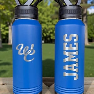 May include: Two blue water bottles with black lids and silver accents. One bottle has the name "JAMES" in silver block letters. The other bottle has a silver "Wb" logo. Both bottles are on a wooden surface.
