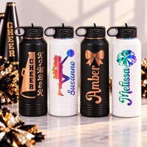 May include: Four cylindrical water bottles with black lids and carrying loops. Two are matte black and two are white. The black bottles feature rose gold graphics of a megaphone with the name Kimber and a bow with the name Amber. The white bottles feature iridescent graphics of a cheerleader with the name Susanne and a pom-pom with the name Melissa.