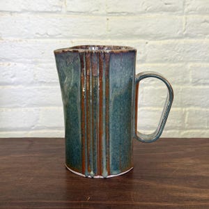 May include: A ceramic pitcher featuring a vertical ribbed design with a glossy glaze in shades of deep teal, blue, and earthy brown. The vessel has a wide pouring spout and a large, curved handle extending from the side, showcasing a reactive finish.
