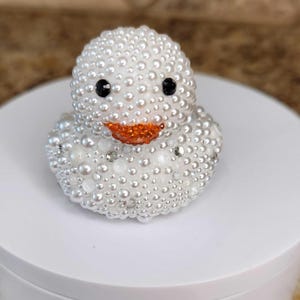 May include: A decorative duck figurine covered in white pearls and rhinestones. The duck has black eyes and an orange beak. The figurine is on a white surface.