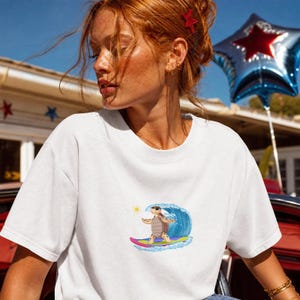 Könnte beinhalten: Ein weißes T-Shirt mit kurzen Ärmeln und einem Grafikdruck einer braunen Schildkröte mit Sonnenbrille, die auf einem pinkfarbenen und violetten Surfbrett vor einer großen blauen Meereswelle und einer kleinen gelben Sonne surft.