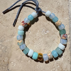 May include: A beaded bracelet featuring a variety of colorful beads in shades of blue, white, and beige, with a gold-colored cylindrical bead. The bracelet is strung on a blue cord with silver-colored metal tips.