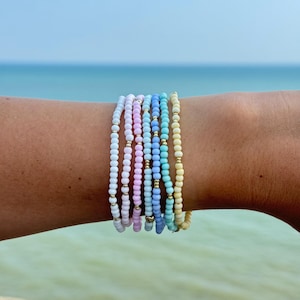 May include: Several beaded bracelets in pastel shades of white, pink, blue, and yellow. Each bracelet is made with small, round beads and gold-coloured spacers. The bracelets are stacked on a wrist, with a blurred ocean backdrop.