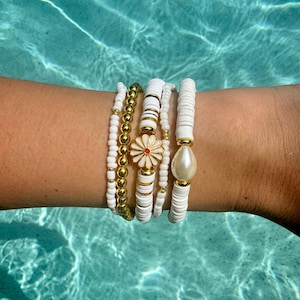 Puede incluir: Una colección de pulseras de cuentas blancas y doradas. Una pulsera presenta un dije de margarita con pétalos rojos y blancos. Otra pulsera tiene una gran perla blanca. Las pulseras están apiladas en una muñeca, con un fondo de agua turquesa.
