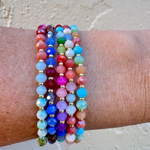 May include: Six beaded bracelets in a spectrum of colours, including red, blue, green, and pink. Each bracelet has faceted beads separated by small gold-coloured spacers. The bracelets are stacked on a wrist.