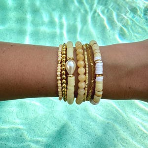 Könnte beinhalten: Eine Sammlung von Armbändern aus Perlen in Gelb-, Creme- und Goldtönen. Die Armbänder weisen verschiedene Designs auf, darunter kleine Perlen, größere runde Perlen und scheibenförmige Perlen. Eine große, birnenförmige Perle ist ein Blickfang auf einem Armband. Die Armbänder sind auf einem Handgelenk gestapelt.