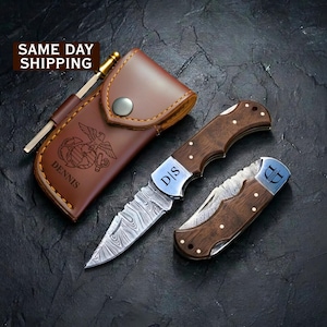 May include: A brown leather pouch with a snap closure and an engraved eagle, globe, and anchor emblem with the name DENNIS, accompanied by a folding pocket knife with a Damascus steel blade and wood handle, and a second folded wood-handled knife.