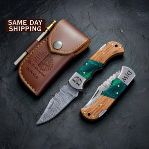 May include: A folding pocket knife with a Damascus steel blade, a wood and green resin handle, and the initials D|W. Beside it is a second open knife with the name DONALD and a brown leather sheath featuring the text MULTIPLE SCLEROSIS.