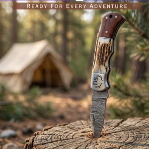 May include: A Damascus steel folding knife with a wooden handle and antler accents is embedded in a tree stump. The knife has a personalized engraving and the text "READY FOR EVERY ADVENTURE" is at the top.