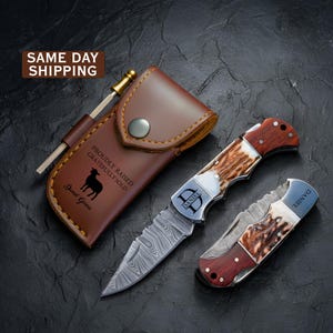 May include: A brown leather sheath with a snap closure, a metal sharpening rod, and two folding pocket knives. The sheath features a silhouette of a sheep and the text PROUDLY RAISED GRATEFULLY SOLD. The knives have wood and antler handles.