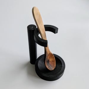 May include: A black spoon rest with a wooden spoon. The spoon rest has a circular base and a curved holder for the spoon. The wooden spoon is light brown and has a rounded bowl. The spoon rest is made of a dark material.