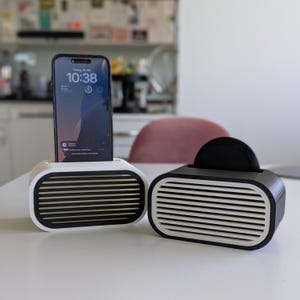 May include: Two modern phone stands. One is white with black and gold horizontal slats, holding a smartphone. The other is black and white, with a black disc in the slot. Both are on a white surface.