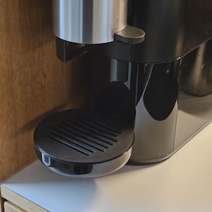 May include: A black coffee machine with a silver top and a drip tray. The coffee machine is sitting on a white worktop.