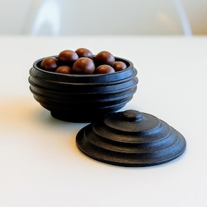 Könnte beinhalten: Eine schwarze, runde Schüssel mit Deckel und geripptem Design. Die Schüssel enthält mehrere braune, kugelförmige Objekte. Der Deckel liegt daneben. Schüssel und Deckel befinden sich auf einer weißen Oberfläche.