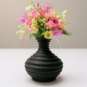 Puede incluir: Un jarrón negro texturizado, con un diseño acanalado, contiene un ramo de flores coloridas. Incluye rosas rosas, flores amarillas y rosas y hojas verdes. El jarrón es un objeto decorativo.
