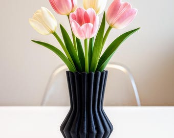 May include: A black, geometric vase holds a bouquet of tulips. The tulips have pink and white petals with green stems and leaves. The vase is made of a dark material and has a ridged design.
