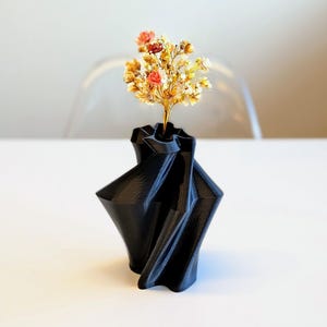 May include: A black, geometric vase with a unique, folded design. The vase holds a small bouquet of dried flowers in shades of yellow, orange, and pink. The vase is made of a solid, dark material and sits on a white surface.