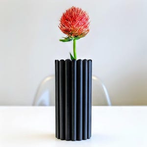 Puede incluir: Un jarrón negro cilíndrico con estrías verticales sostiene una vibrante flor roja y naranja. El jarrón tiene un borde superior festoneado y se apoya sobre una superficie blanca. El tallo verde de la flor contrasta con el jarrón oscuro.