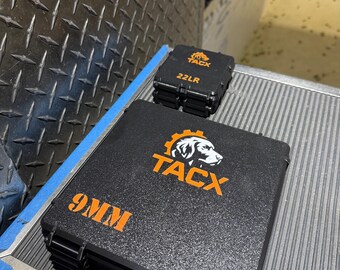 Könnte beinhalten: Zwei schwarze Koffer mit orangefarbenem Text und Logos. Der größere Koffer trägt die Aufschrift "9MM" und zeigt das "TACX"-Logo mit einer Hundesilhouette. Ein kleinerer Koffer trägt die Aufschrift "22LR" und hat ebenfalls das "TACX"-Logo.
