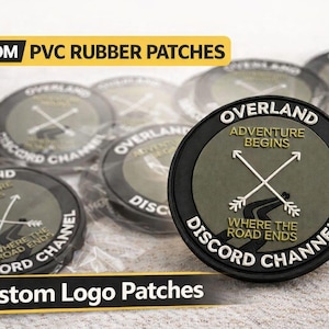 Op de afbeelding: Op maat gemaakte PVC rubberen patches met een cirkelvormig ontwerp. De patches tonen de tekst "OVERLAND ADVENTURE BEGINS" en "DISCORD CHANNEL" rond een gekruiste pijlgrafiek. De patches zijn zwart, olijfgroen en wit.