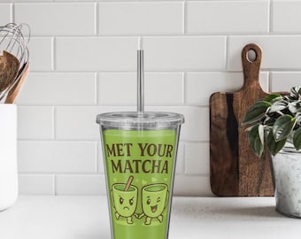 Puede incluir: Un vaso de plástico transparente lleno de líquido verde, con una pajita y tapa plateadas. El vaso tiene el texto "MET YOUR MATCHA" y un gráfico de dibujos animados de dos tazas de matcha. El fondo incluye una pared de azulejos blancos y una tabla de cortar de madera.