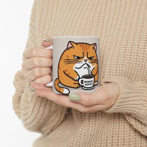 Puede incluir: Taza de cerámica blanca con un gato naranja de dibujos animados sosteniendo una taza de café que dice "¿Lunes otra vez?"