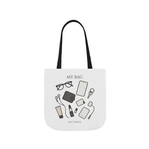 Puede incluir: Bolso tote blanco con asas negras. El bolso presenta un gráfico de objetos cotidianos, que incluyen gafas, un teléfono, una cartera, llaves, lápiz labial y otros accesorios. El texto en el bolso dice "MY BAG" y "MY THINGS."