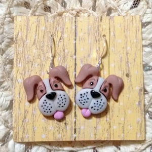 May include: A pair of dangle earrings featuring a cartoon dog face with brown ears, white fur, black eyes, a black nose, and a pink tongue. The earrings are made of polymer clay and have silver hooks.