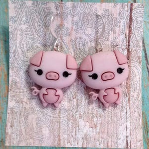 May include: A pair of pink pig earrings with black eyes and a curly tail. The earrings are made of a smooth material and have silver-colored ear wires. The earrings are displayed on a patterned background.