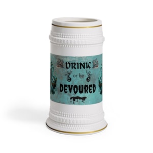 Pode incluir: Caneca de cerveja de cerâmica branca com detalhes dourados e um rótulo turquesa. O rótulo diz "DRINK or be DEVOURD" em letras góticas pretas. A caneca tem um design texturizado.