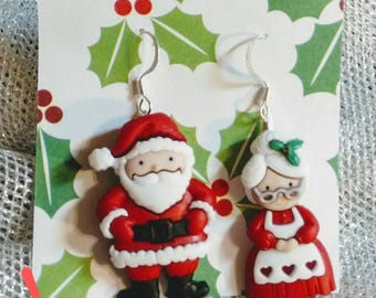 May include: A pair of Christmas-themed earrings featuring Santa Claus and Mrs. Claus. Santa is wearing a red suit with white trim and a white beard. Mrs. Claus is wearing a red dress with a white apron and a white bonnet.