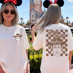 Puede incluir: Camiseta de color crema con un gr&aacute;fico del Sheriff Woody en la parte delantera y trasera. El dise&ntilde;o trasero incluye el texto "Sheriff Woody" sobre un patr&oacute;n de cuadros marrones y blancos. La camiseta se combina con pantalones cortos a cuadros rojos y blancos.
