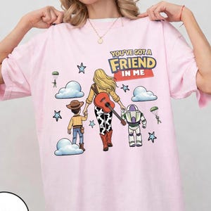 May include: A light pink t-shirt featuring a graphic of a cowboy, a cowgirl with a guitar, and a space ranger character. The text reads YOU'VE GOT A FRIEND IN ME. The design includes blue clouds, stars, and parachuting green soldiers on a light pink background.
