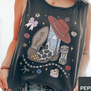 Puede incluir: Una camiseta sin mangas de color gris carb&oacute;n con un estampado gr&aacute;fico de una bota de vaquero con manchas de vaca, un sombrero de vaquero rojo, un lazo rosa y bloques de letras que forman T-S. El dise&ntilde;o incluye estrellas y flores.