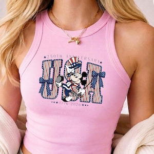 Op de afbeelding: Een lichtroze geribbelde tanktop met een afbeelding van Mickey Mouse in een patriottische hoed en outfit. De tekst luidt 250TH ANNIVERSARY USA 1776 2026, met rood-wit gestreepte letters en blauwe strikken met witte sterren aan de zijkanten.