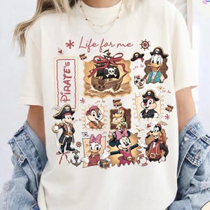 May include: Ivory-colored t-shirt featuring a pirate-themed design with cartoon characters. The design includes a pirate ship, skull, and various characters in pirate attire. The text "Life for me" and "Pirate's" are also visible.