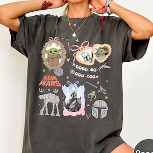 Puede incluir: Camiseta de color pimienta con gr&aacute;ficos de Star Wars. El dise&ntilde;o incluye im&aacute;genes de Grogu, el Mandaloriano y un caminante AT-AT. El texto "Star Wars" se muestra de forma destacada, junto con otros elementos decorativos.