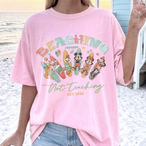 May include: A pink t-shirt with the words "BEACHING Not Teaching" in colorful letters and a graphic of cartoon characters on surfboards. The shirt also has the text "#Retired EST. 2026".