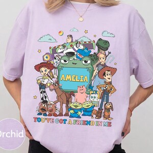 May include: A light purple short-sleeve t-shirt featuring a colorful graphic of various animated toy characters surrounding a green frog-shaped frame with the name AMELIA printed inside and the text YOU'VE GOT A FRIEND IN ME below.