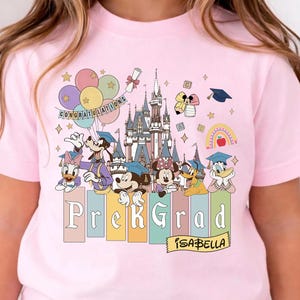 May include: Pink t-shirt featuring a colorful graphic of Disney characters celebrating a Pre-K graduation. The design includes a castle, balloons, and the text "Prek Grad" with the name "Isabella".