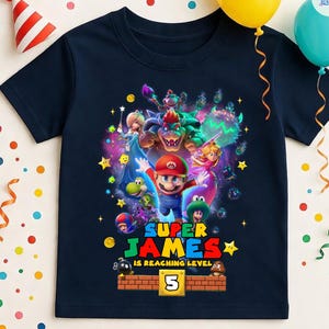 May include: Navy blue t-shirt featuring a colorful graphic of Super Mario characters. The design includes the text "SUPER JAMES IS REACHING LEVEL 5" with a brick-style border. Party decorations, including balloons and a party hat, are in the background.