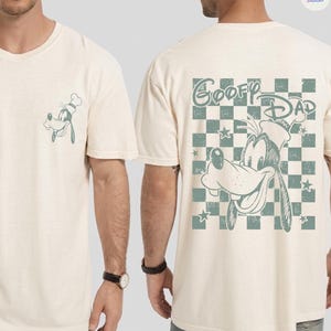 May include: Cream-colored t-shirt featuring a Goofy graphic on the front pocket and back. The back design includes the text "Goofy Dad" above a checkered pattern with stars. The shirt is a casual wear item.