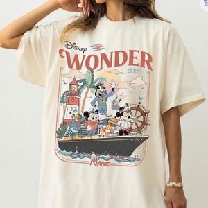 Puede incluir: Camiseta color crema con un gr&aacute;fico del crucero Disney Wonder con Mickey Mouse, Donald Duck y otros personajes. El dise&ntilde;o incluye el texto "Disney Wonder 2026" en rojo y un faro.