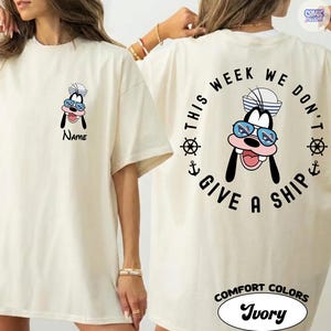 May include: Ivory-colored t-shirt featuring a cartoon character wearing a sailor hat and sunglasses. The front of the shirt has the character's face and the word "Name". The back of the shirt has the phrase "This week we don't give a ship" in a circular design.