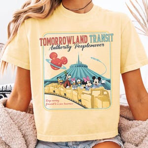 Puede incluir: Camiseta amarilla clara con un dise&ntilde;o gr&aacute;fico retro del paseo Tomorrowland Transit Authority Peoplemover, con Mickey y Minnie Mouse. El dise&ntilde;o incluye el texto "Tomorrowland Transit Authority Peoplemover" y "Keep moving forward to a new tomorrow."
