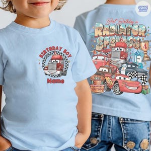 May include: Light blue t-shirt featuring a cartoon truck with the text "Birthday Boy" and "Name" on the front. The back of the shirt displays a colorful graphic with the text "Best Birthday in Radiator Springs" and various cartoon cars.