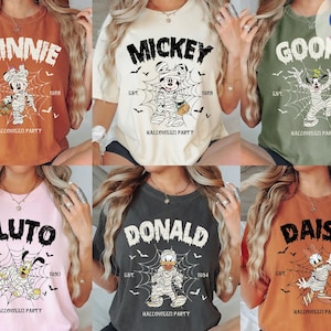 May include: A collection of six short-sleeve t-shirts in various colors, each featuring a Disney character dressed as a mummy in a spiderweb, with the text "HALLOWEEN PARTY" and the character's name printed above, plus "EST." and a year below.
